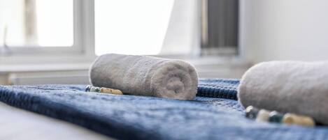Ein gerolltes Handtuch auf einem blauen, strukturierten Stoff, möglicherweise eine Badmatte, mit einem Fenster im Hintergrund.