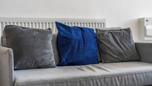 Ein graues Sofa mit drei Kissen, eines blau und zwei grau, vor einer weißen Heizung.
