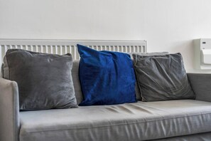 Ein graues Sofa mit drei Kissen, eines blau und zwei grau, vor einer weißen Heizung.