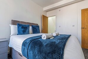 Una cama bien hecha con una colcha y almohadas azules, una mesita de noche y una puerta de madera al fondo.