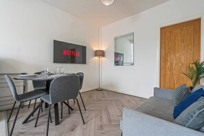 A modern living room with a grey sofa, a dining area with a table and chairs, a wall-mounted TV displaying 'NETFLIX', and a wooden door. - 2 Bed Duplex | Jewellery Quarter | Sleeps 4 (West Midlands)