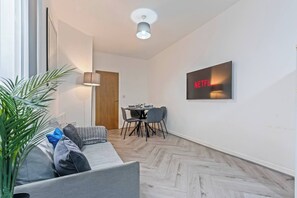 A modern living room with a sofa, a dining table with chairs, a lamp, and a wall-mounted TV displaying the Netflix logo. - 1 Bed Flat | Jewellery Quarter | Sleeps 2 (West Midlands)