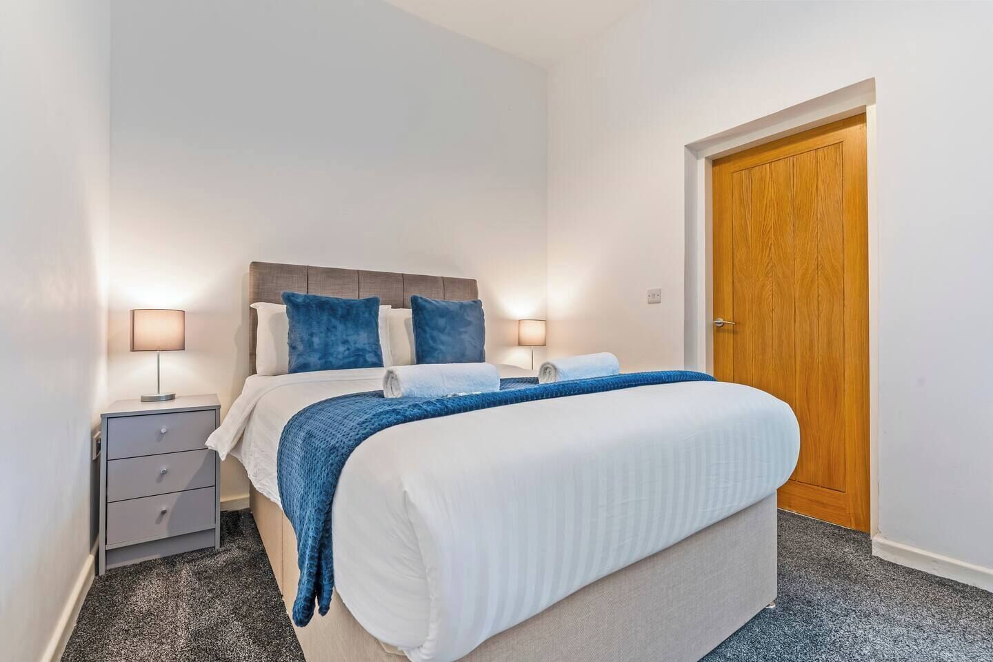 A bedroom with a bed, two bedside tables, a wooden door, and a carpeted floor.