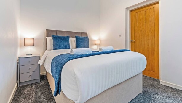 Une chambre à coucher avec un lit, deux tables de chevet, une porte en bois et un sol recouvert d’une moquette.