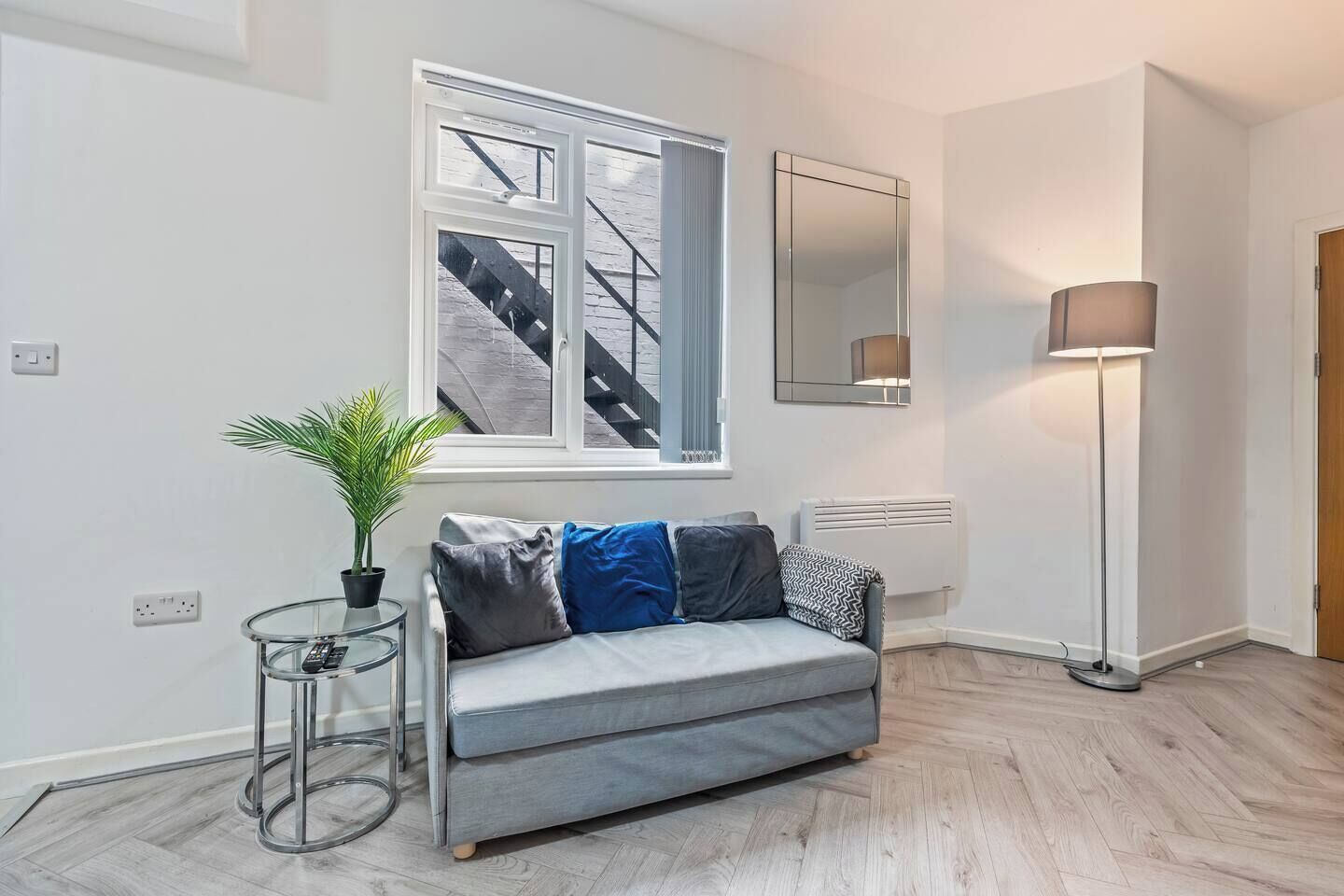A modern living room with a grey sofa, a small side table with a plant, a floor lamp, and a large window with a view of a staircase.