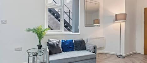 Una sala de estar moderna con un sofá gris, una mesita auxiliar con una planta, una lámpara de pie y un ventanal con vista a una escalera.