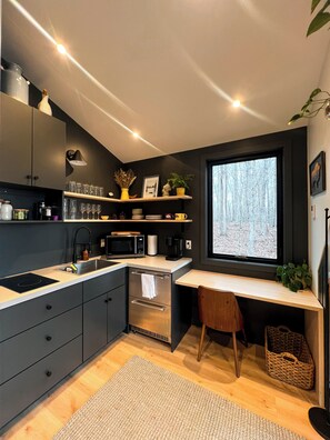 A modern kitchen with a built-in oven, microwave, and sink. There is a window with a view of trees, and a small desk with a chair. - Bison Farm - Cabin "Brisa" (Culpeper)