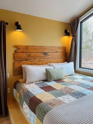 A bedroom with a wooden headboard, a quilted bedspread, and two wall-mounted lamps. - Bison Farm - Cabin "Brisa" (Culpeper)