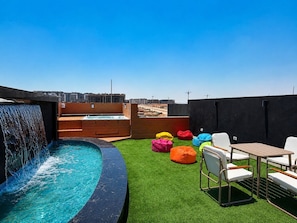 A rooftop pool with a waterfall feature, surrounded by green artificial turf and colorful bean bags.