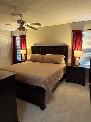 A bedroom with a bed, a nightstand, a lamp, a ceiling fan, and a window with curtains. - 2-Bedroom Condo in Starkville - Perfect for Game Days & Getaways!! (Starkville)