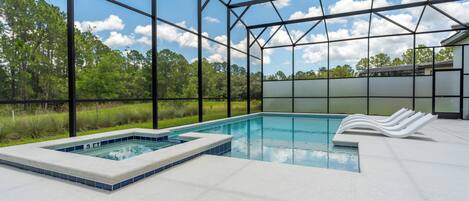A swimming pool with a hot tub, surrounded by a covered area with a metal frame and glass walls, set against a backdrop of trees and a clear sky.