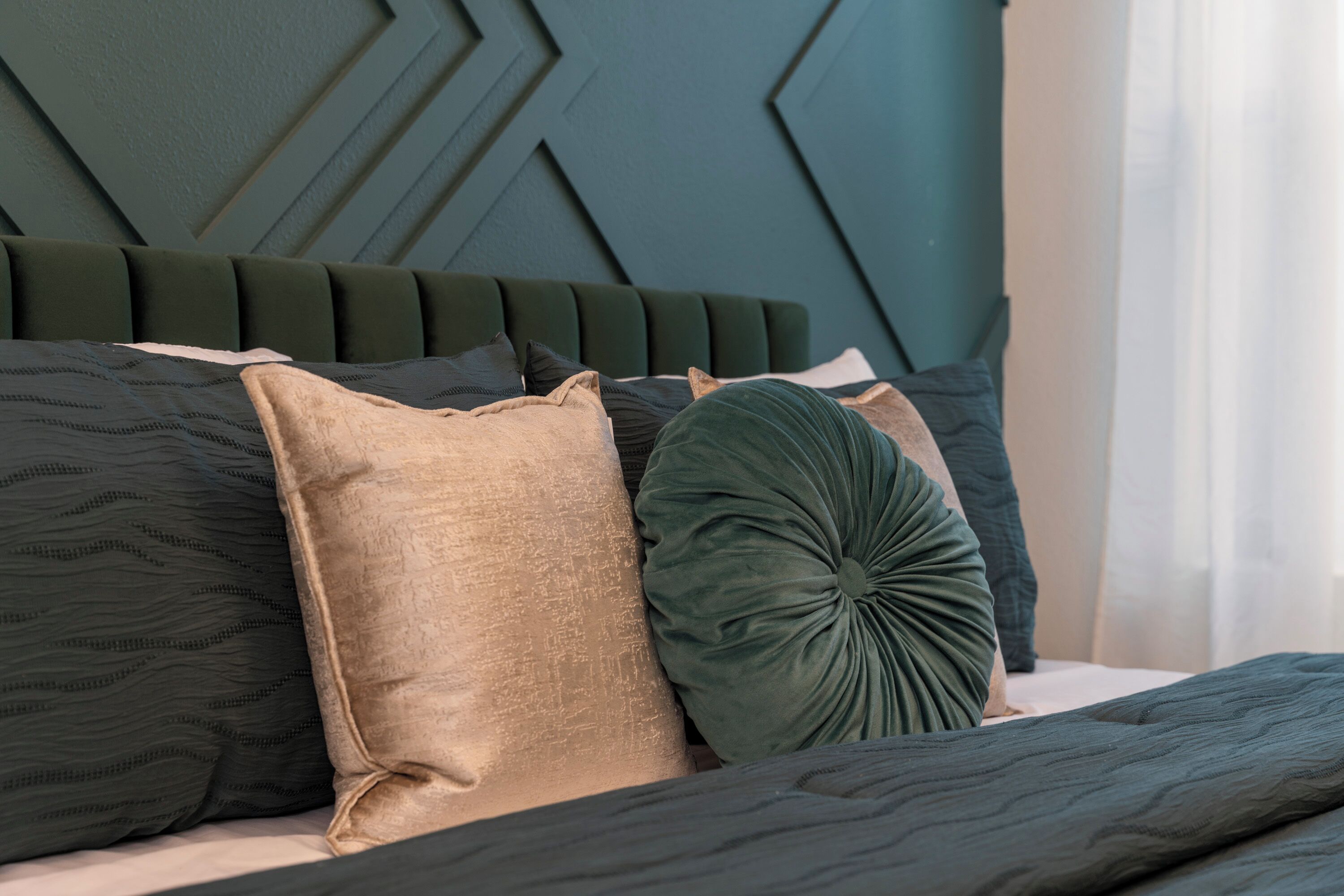 Un lit avec une tête de lit vert foncé, plusieurs oreillers de différentes textures et couleurs, et une couvre-lit à motifs.