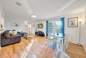 A spacious living area with a sofa, coffee table, and a dining table with chairs. There is a staircase leading to another level, and a large window with curtains. - Sweetwater Cove:  3-Level Lakefront Retreat (Moneta)