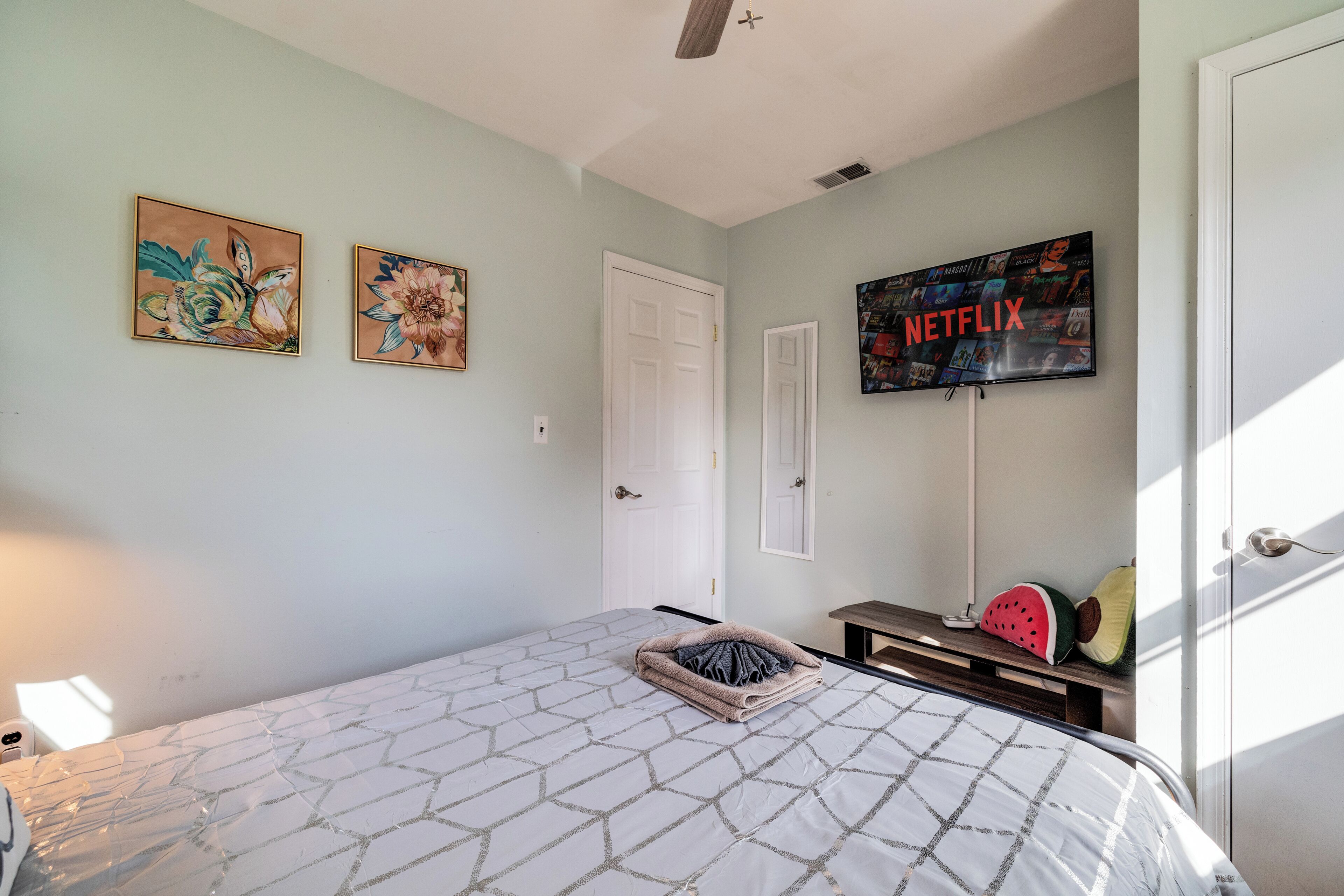 Un dormitorio con una cama, un televisor montado en la pared y dos cuadros enmarcados.