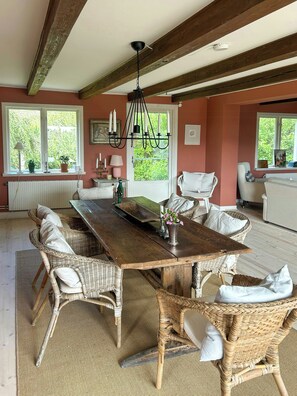 A dining area with a wooden table surrounded by wicker chairs, a chandelier, and a view of greenery outside. - Farmhouse with view towards Kullaberg (BÅSTAD)