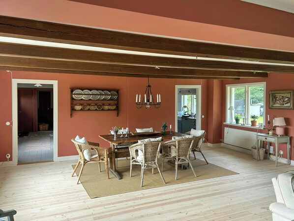 A dining area with a wooden table and wicker chairs, a chandelier, and a view of the kitchen through an open doorway. - Farmhouse with view towards Kullaberg (BÅSTAD)