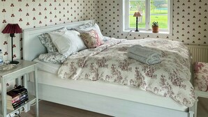 Een slaapkamer met een bed, een nachtkastje met boeken, een lamp en een raam met uitzicht op groen.