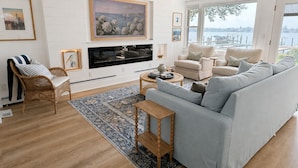 A modern living room with a large light grey sofa, a wooden coffee table, and a TV mounted on the wall. There is a large window providing a view of the outdoors.
