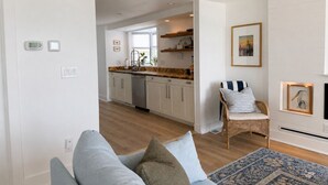 A modern kitchen with white cabinets, a dishwasher, and a window. There is a wicker chair with cushions and a blue patterned rug.