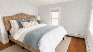 A bedroom with a woven headboard, a bed with multiple pillows, a bedside lamp, and a window with blinds.