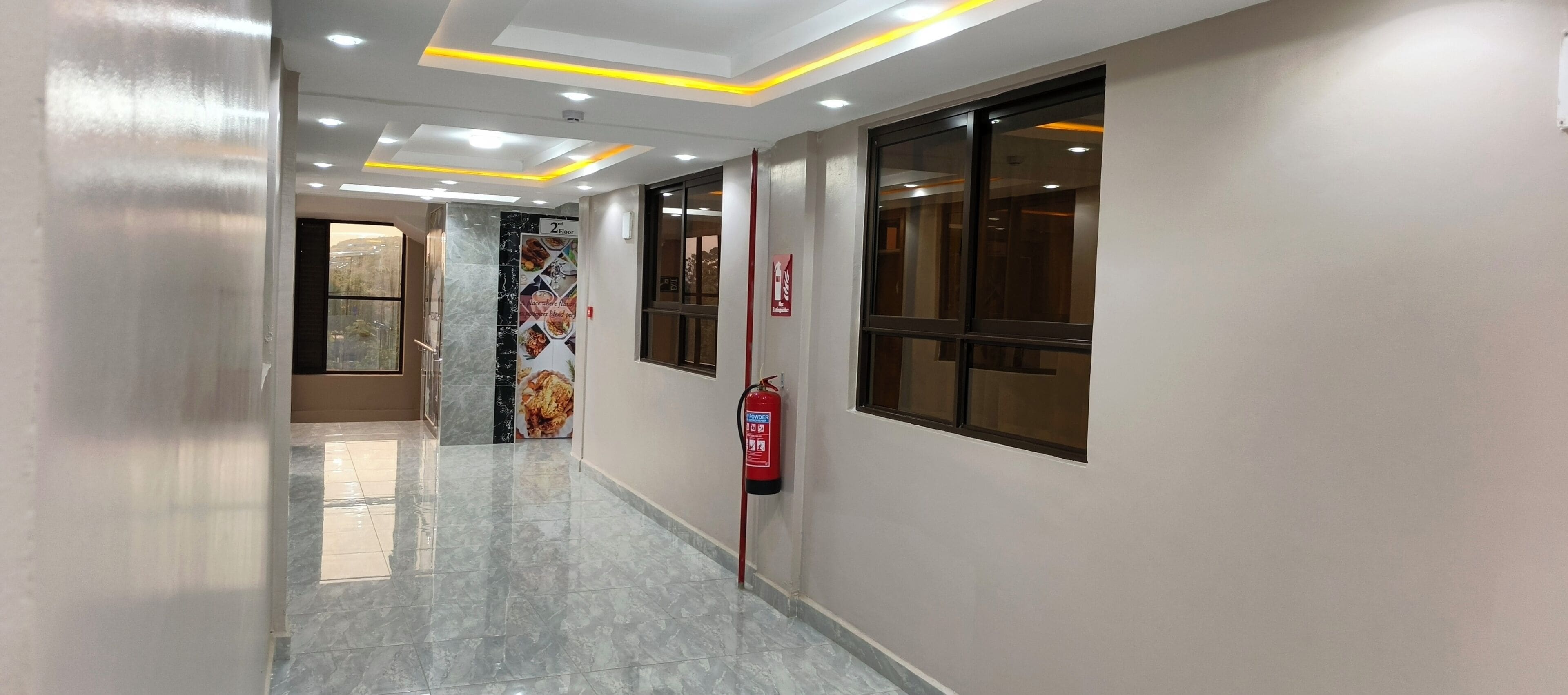Corredor de hotel com um extintor preso na parede, um sinal de saída de incêndio e uma janela com vista.