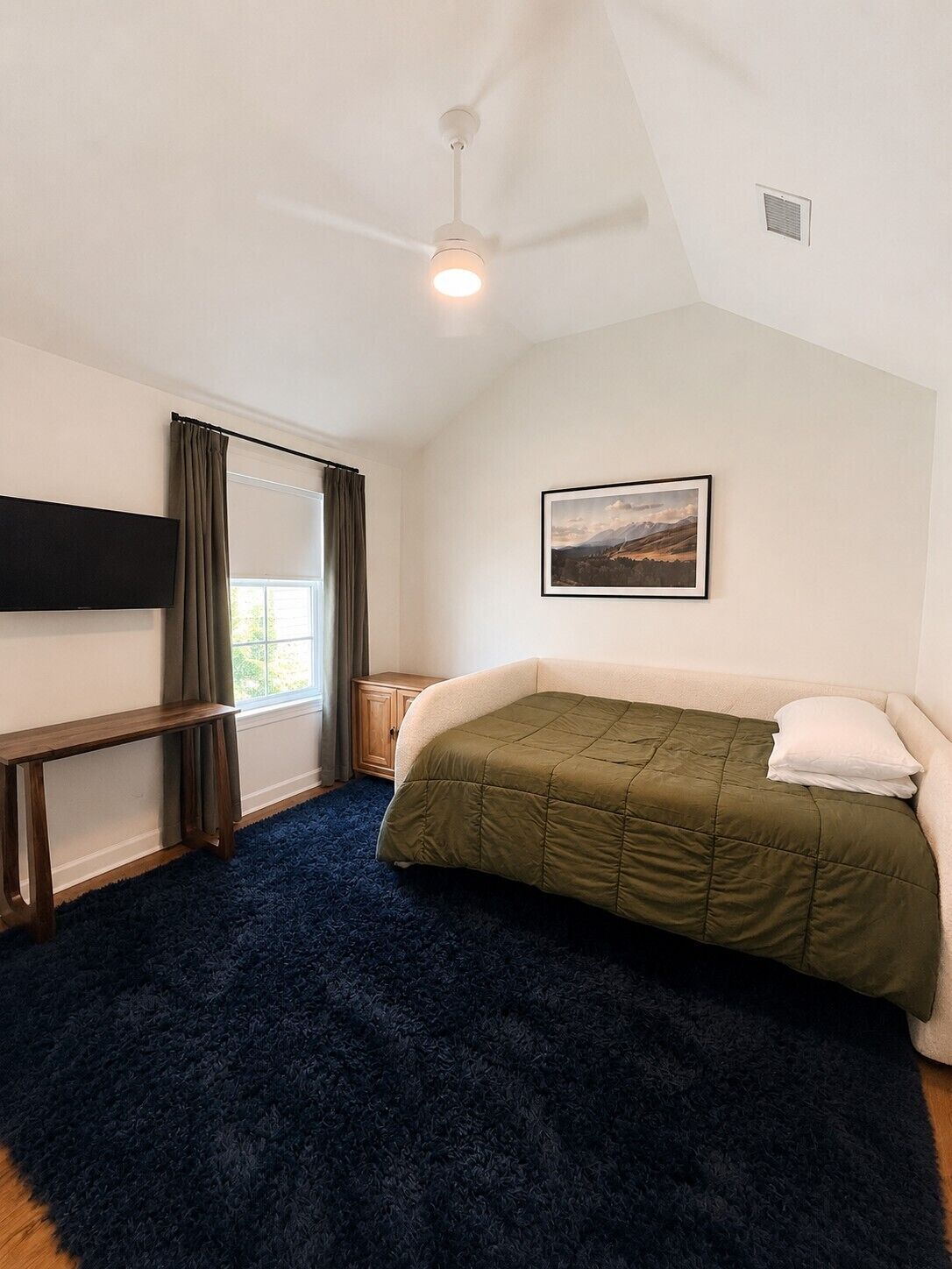 A bedroom with a bed, a desk, a chair, a window with curtains, a ceiling fan, and a picture on the wall.