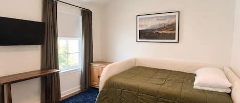 A bedroom with a bed, a desk, a chair, a window with curtains, a ceiling fan, and a picture on the wall.