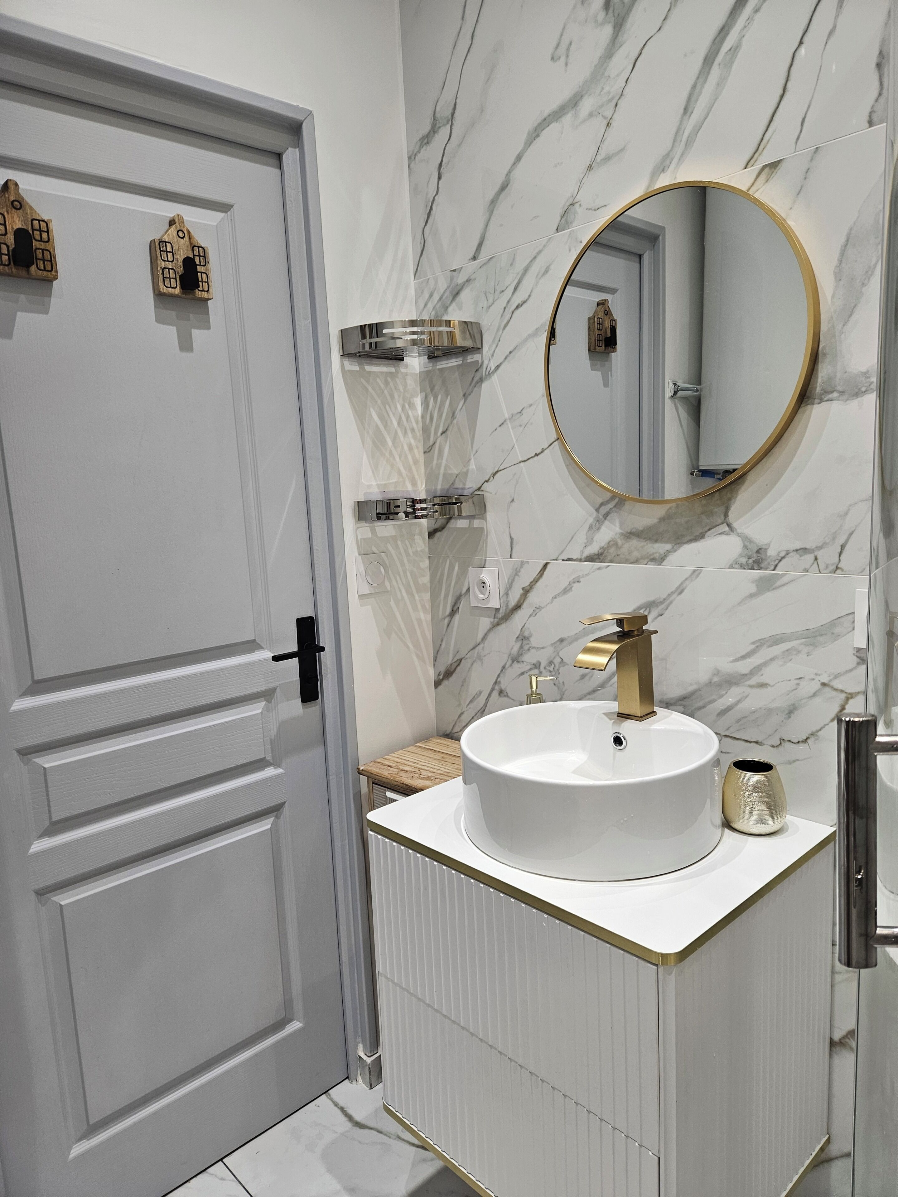 A bathroom with a white door, a round mirror with a gold frame, a white sink with a gold faucet, and a marble wall.