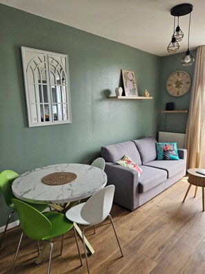 A living room with a round marble table, green chairs, a grey sofa with decorative pillows, and a wall clock. - Renovated and well equipped apartment in the heart of Mers and close to the beach. (Mers-les-Bains)