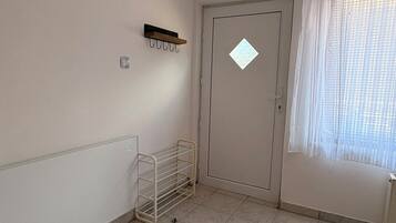 Ein Zimmer mit Fliesenboden, Heizkörper, einer Tür mit einem rautenförmigen Fenster und einem Fenster mit durchsichtigen Vorhängen.