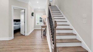 A hallway with a wooden staircase, a bed in the adjacent room, and a window with blinds. - Hallandale Hideaway, luxury townhome, sleeps 8 with POOL. (Murrells Inlet)
