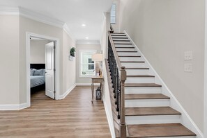 A hallway with a wooden staircase, a bed in the adjacent room, and a window with blinds. - Hallandale Hideaway, luxury townhome, sleeps 8 with POOL. (Murrells Inlet)