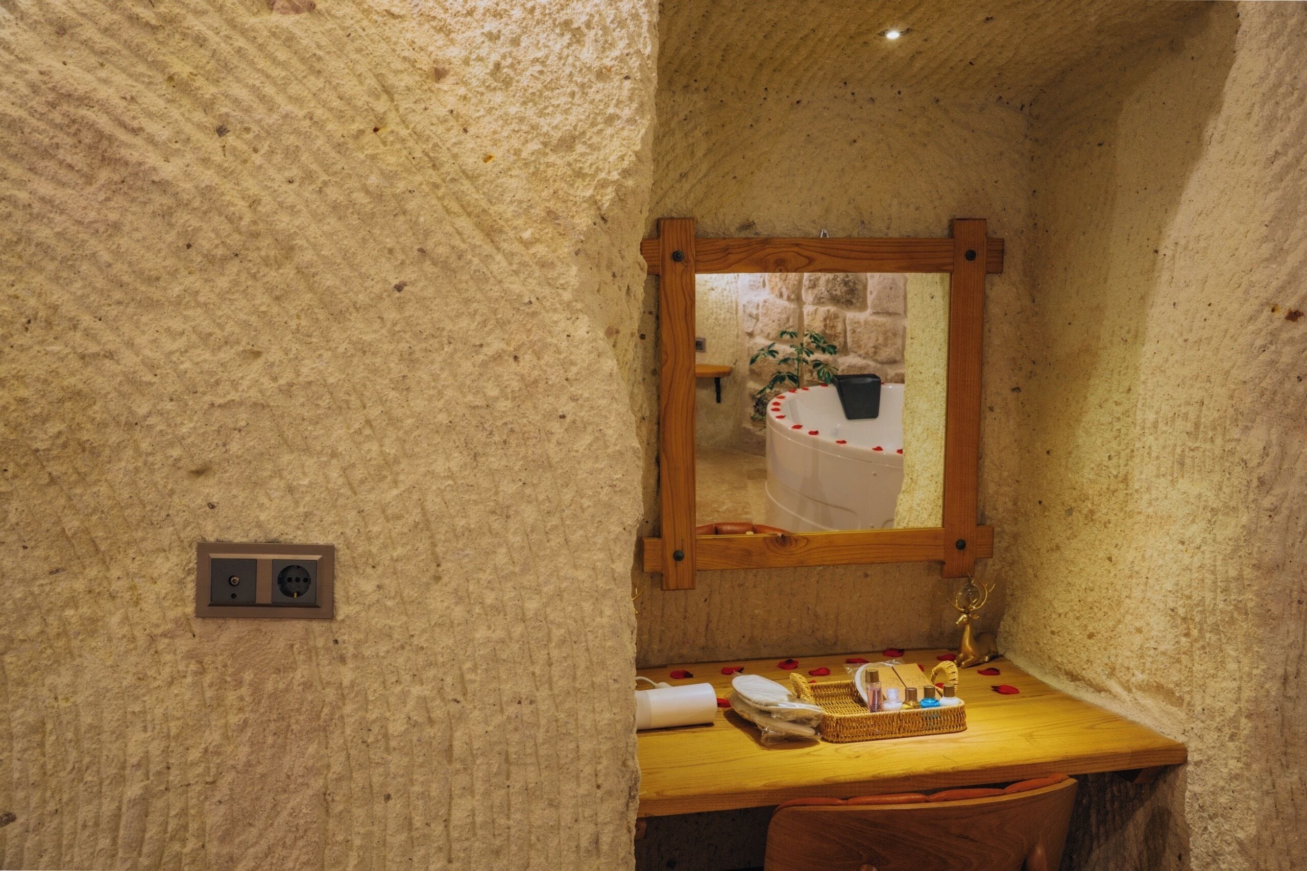 A rustic bathroom with a stone wall, a wooden-framed mirror, a sink with toiletries, and a small plant.