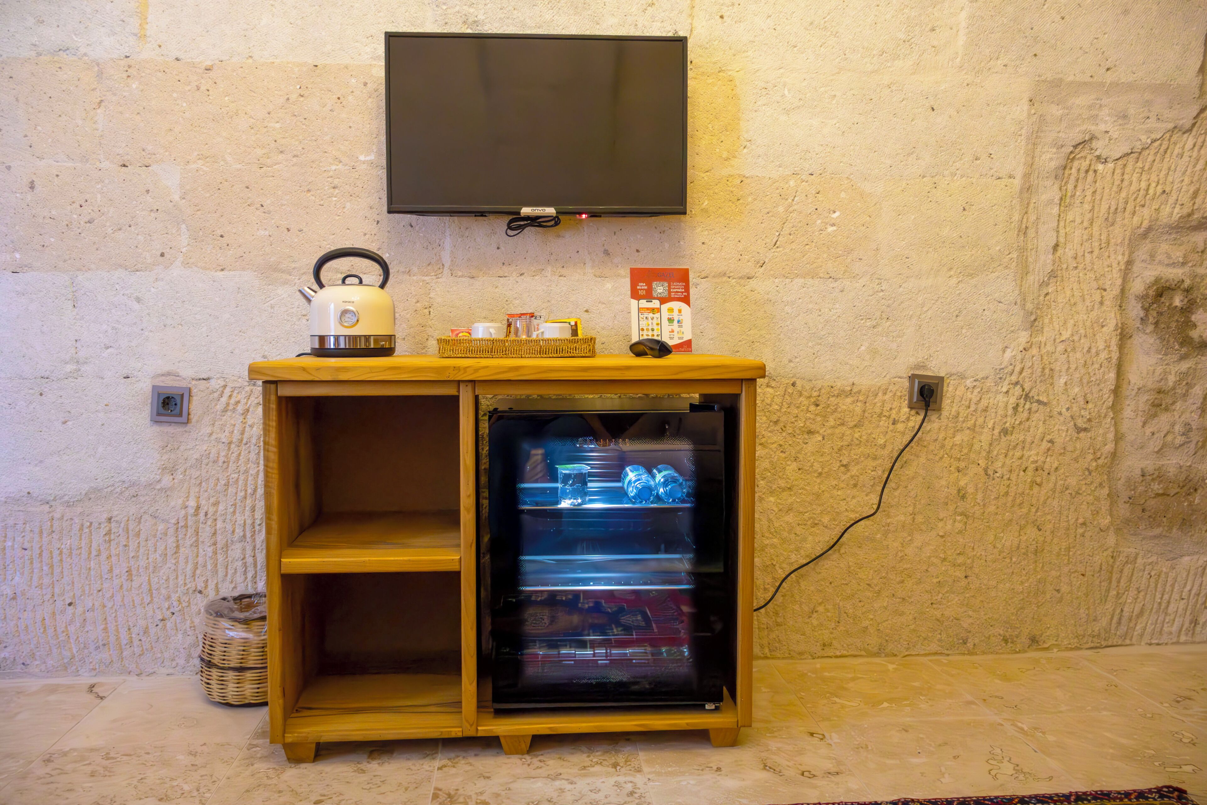 Ein kleines Holzschränkchen mit einem darüber angebrachten Fernseher, einem Wasserkocher und einem kleinen Kühlschrank mit Glastür.
