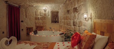 A stone-walled room with a large bathtub, a bed with rose petals, and a red curtain.