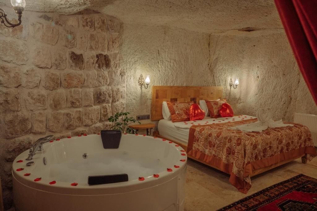 A room with a stone wall, a wooden bed, a large bathtub with rose petals, and a red curtain.