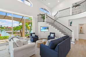 Uma sala de estar espaçosa com uma janela grande que dá para uma piscina, uma escada com corrimão preto e uma televisão de tela plana na parede.