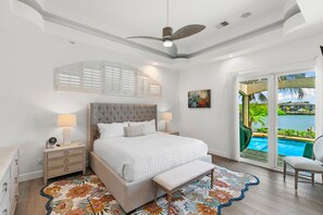 Quarto com uma cama grande, ventilador de teto, mesa de cabeceira com luminária, um banquinho e vista para uma piscina através de uma porta de vidro.