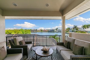 A covered outdoor seating area with wicker furniture, a round table, and a view of a body of water and buildings in the distance. - Waterfront Marco Island Home w/ Pool, Spa & Kayaks (Marco Island)