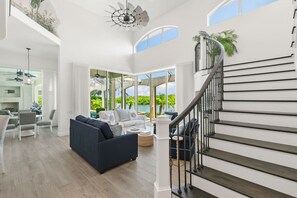 A spacious living area with a blue sofa, white armchairs, and a dining table. A staircase with a black railing leads to an upper level. - Waterfront Marco Island Home w/ Pool, Spa & Kayaks (Marco Island)