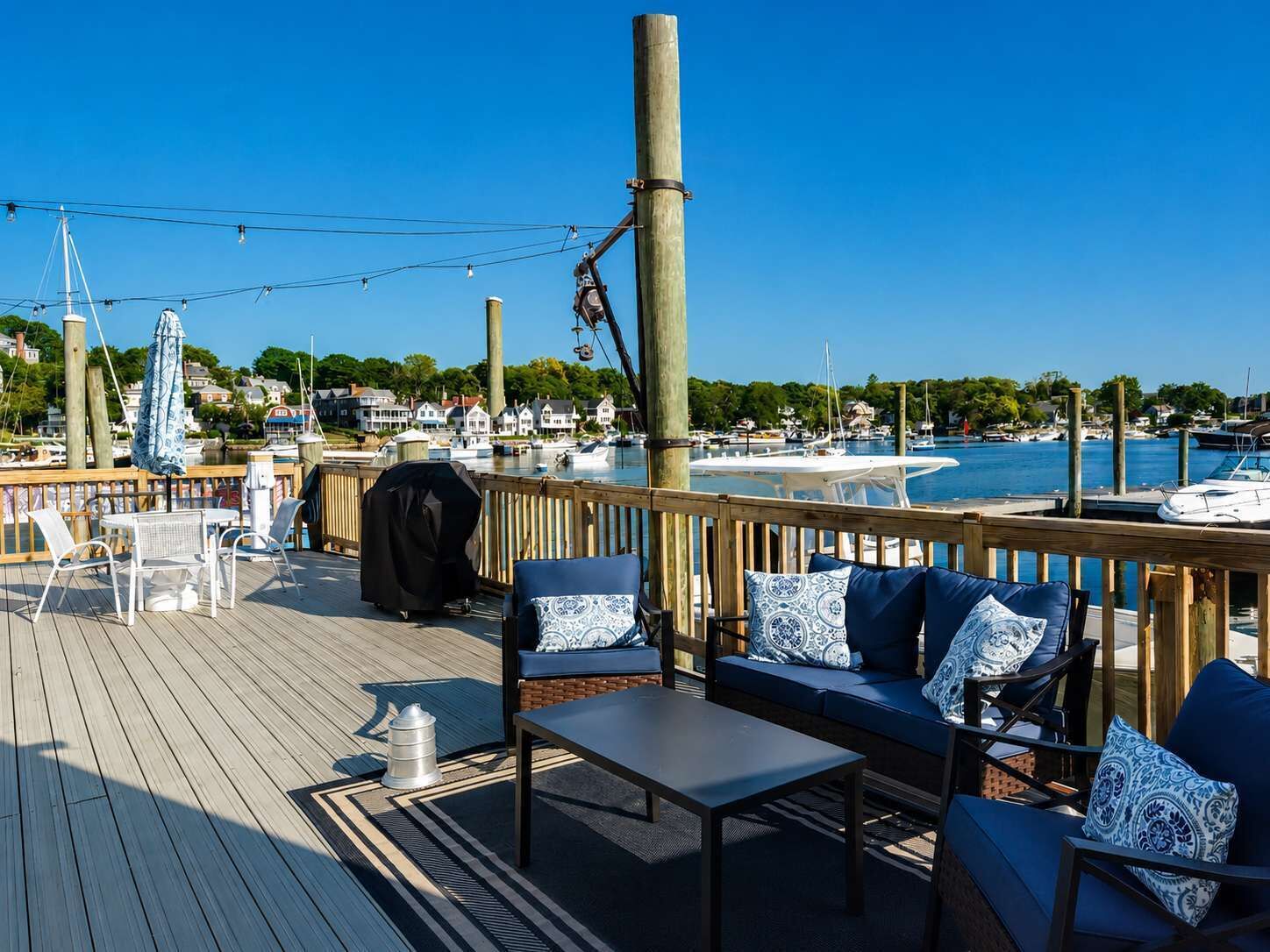 A wooden deck with outdoor seating, a grill, and a view of a marina with boats and houses in the background.