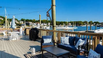 A wooden deck with outdoor seating, a grill, and a view of a marina with boats and houses in the background.