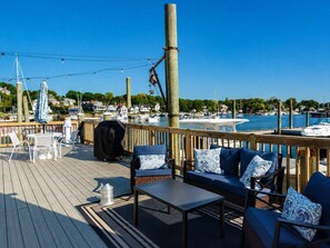 A wooden deck with outdoor seating, a grill, and a view of a marina with boats and houses in the background.