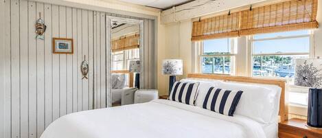 A bedroom with a large bed, a wooden headboard, a window with a view of boats, and a wall with nautical-themed decorations.
