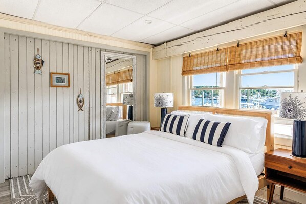 A bedroom with a large bed, a wooden headboard, a window with a view of boats, and a wall with nautical-themed decorations.