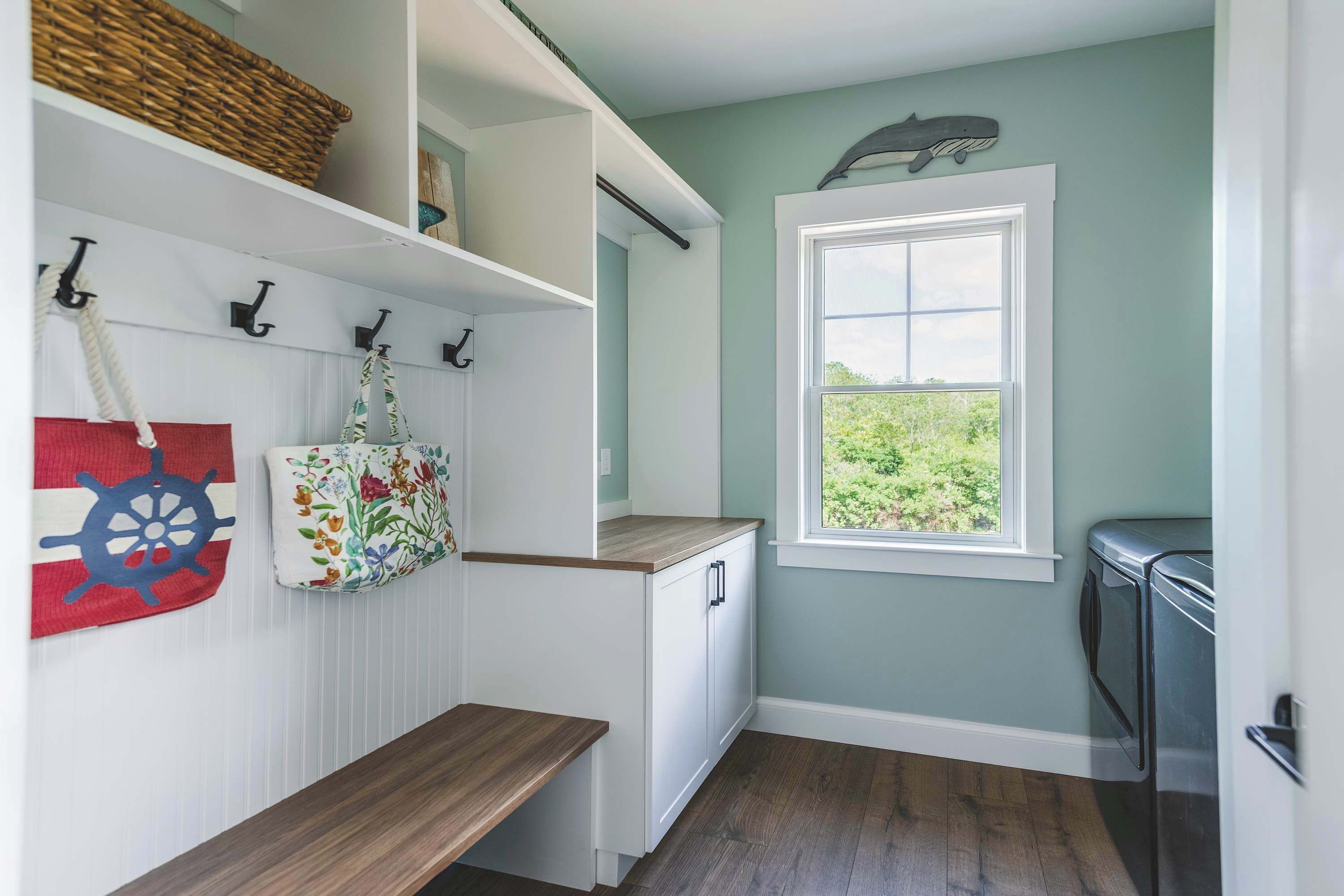 A laundry room with a wooden bench, a window with a view of greenery, and a wall-mounted basket.
