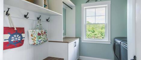 A laundry room with a wooden bench, a window with a view of greenery, and a wall-mounted basket.