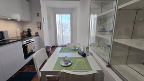 Eine kompakte Küche mit Mikrowelle, Toaster und kleinem Kühlschrank. Ein Esstisch mit zwei Stühlen und eine Vitrine mit Dekorationen sind ebenfalls vorhanden.