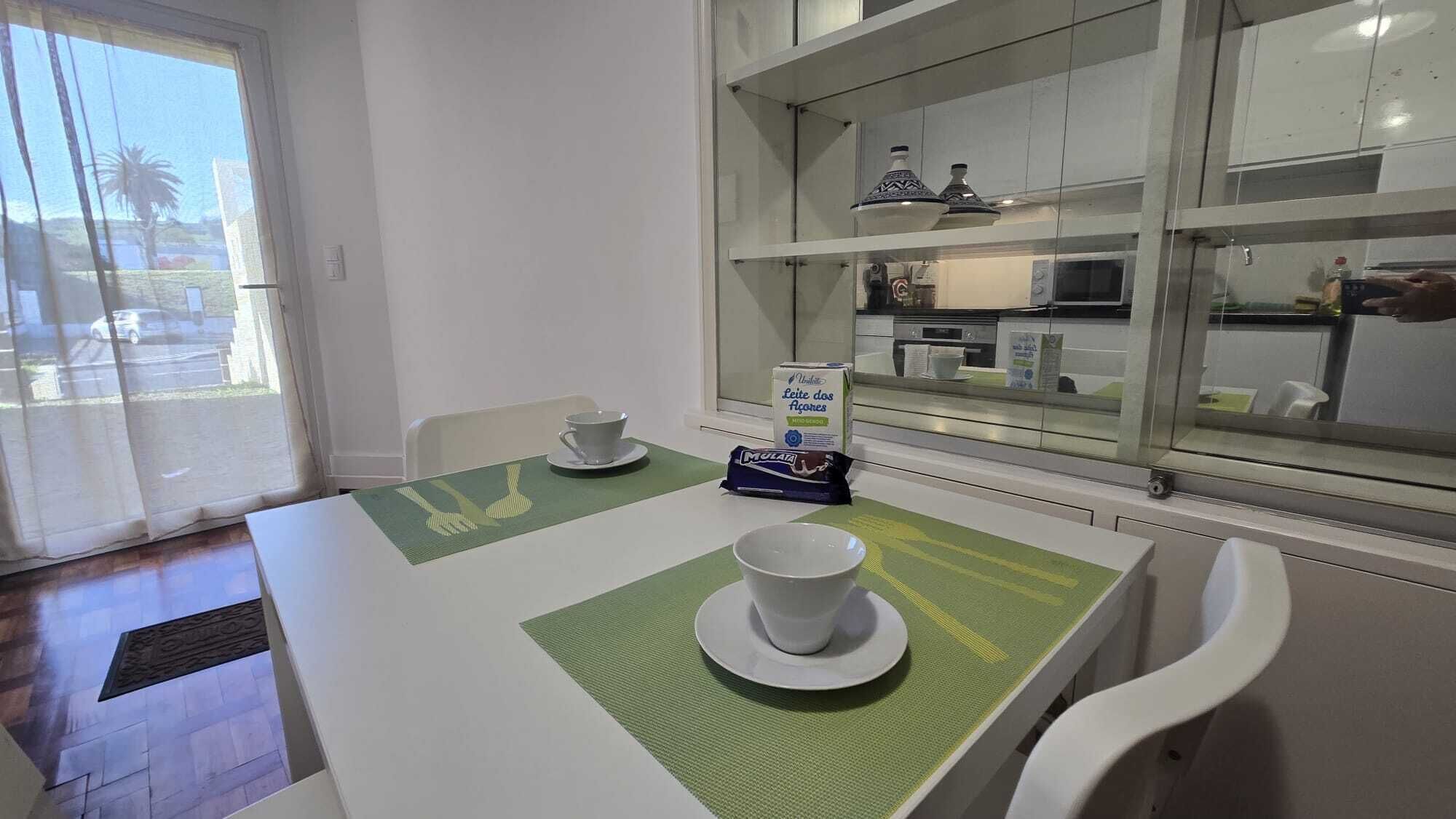 A dining area with a table set for two, a kitchen with glass cabinets, and a view of the outside through a window.