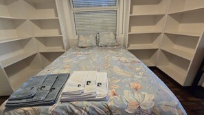 Ein Bett mit floralem Überwurf, ein Fenster mit Jalousien und ein Schrank mit Regalen.